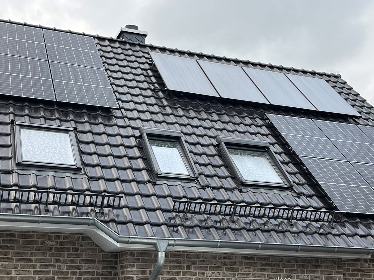 Dach mit Solarpanelen, dunkle Ziegel, drei Fenster, bewölkter Himmel.
